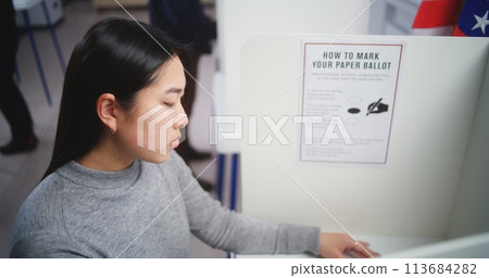 Asian woman comes to voting booth and fills out paper bulletin Asian woman comes to voting booth and fills out paper bulletin 113684282