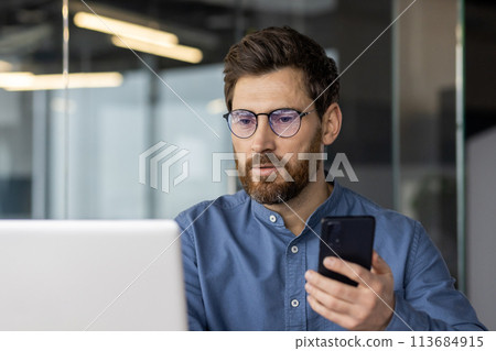 Close-up photo of a serious and focused man in glasses and a shirt working in the office on a laptop, and holding a mobile phone. 113684915