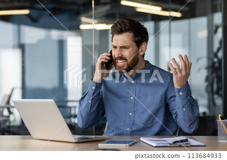 Angry and irritated young businessman sitting in the office at the desk and talking on the phone while waving his hands. 113684933