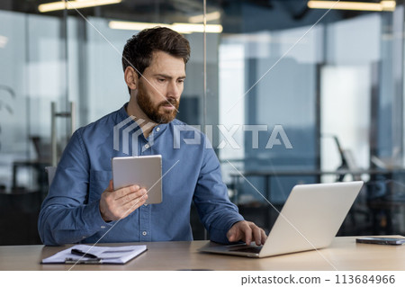 A concentrated and serious young man is sitting at a desk in the office, holding a tablet and working on a laptop. A concentrated and serious young man is sitting at a desk in the office, holding a tablet and working on a laptop. 113684966