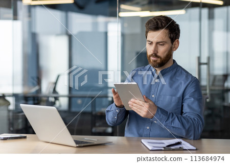 A serious young man is sitting in a modern office in a blue shirt at his desk and using a tablet. A serious young man is sitting in a modern office in a blue shirt at his desk and using a tablet. 113684974