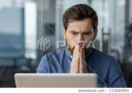 Shocked and worried young male businessman sitting in the office at his desk and looking thoughtfully at the laptop screen with his hands folded together. Shocked and worried young male businessman sitting in the office at his desk and looking thoughtfully at the laptop screen with his hands folded together. 113684983