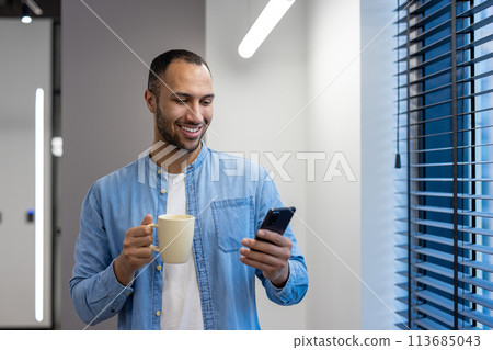 Smiling young arab man working in office, standing by window on work break, holding cup with drink and using mobile phone. Smiling young arab man working in office, standing by window on work break, holding cup with drink and using mobile phone. 113685043