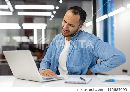 Suffering from pain tired hispanic young pencil sitting in modern office at desk and holding hand behind back. 113685093