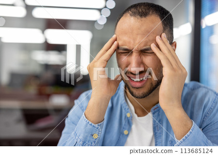 A close-up photo of a Muslim young man sitting in the office behind the workplace and hunched over from anger and tension, holding his head in his hands. A close-up photo of a Muslim young man sitting in the office behind the workplace and hunched over from anger and tension, holding his head in his hands. 113685124