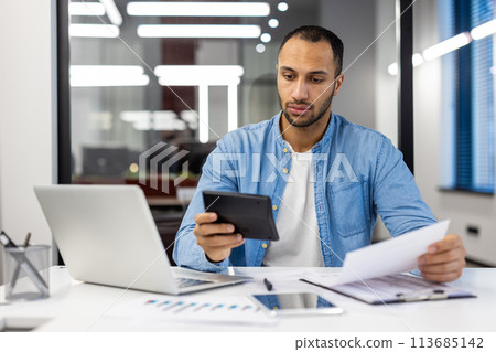 Serious young hispanic male accountant and auditor sitting in office at desk working with documents, bills and calculator. 113685142