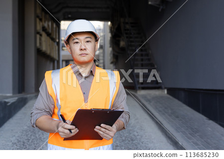 Portrait of serious and successful young Asian man in hard hat and vest standing outside building with folder and pen in hands, inspecting, developing project. Portrait of serious and successful young Asian man in hard hat and vest standing outside building with folder and pen in hands, inspecting, developing project. 113685230