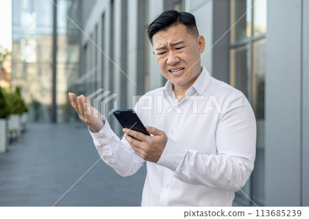 Angry and worried young Asian male businessman standing outside a building, looking upset at the phone screen and throwing up his hands. Angry and worried young Asian male businessman standing outside a building, looking upset at the phone screen and throwing up his hands. 113685239