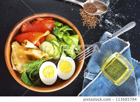 Delicious healthy brunch - boiled egg, smoked salmon, green salad and savory pancakes on a dark background, top view 113685784