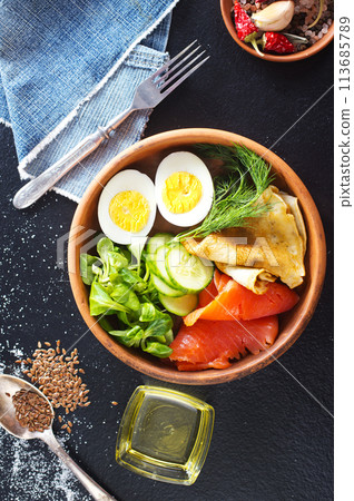 Delicious healthy brunch - boiled egg, smoked salmon, green salad and savory pancakes on a dark background, top view Delicious healthy brunch - boiled egg, smoked salmon, green salad and savory pancakes on a dark background, top view 113685789