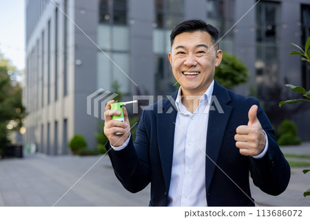 Portrait of a young Asian man in a business suit sitting on the street near the office, holding a cough spray in his hand and showing a super gesture to the camera. 113686072