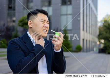 Asian young businessman sitting near working office outside, holding throat with hand, feeling severe pain and using medical spray. 113686076