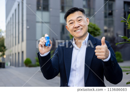 Portrait of a smiling young Asian man in a business suit sitting on a bench near an office building, holding an inhaler in his hand and showing the super sign with his finger to the camera. 113686083