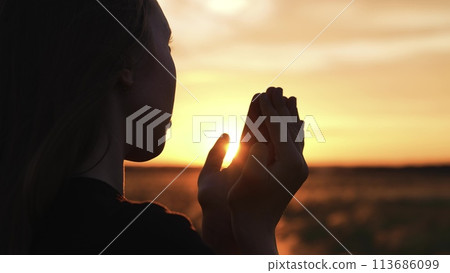 Woman praying with folded hands at sunset silhouette. Religious pious orthodox christian spiritual believing faithful girl at morning dawn sunrise reading prayer bible turning to god jesus christ. 113686099