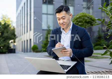 Asian young businessman working outside office center, sitting on bench wearing headphones, holding laptop and making notes in notebook. Asian young businessman working outside office center, sitting on bench wearing headphones, holding laptop and making notes in notebook. 113686113