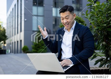 A happy young Asian male businessman is sitting outside an office building and smilingly looking at the screen of a laptop on his lap. A happy young Asian male businessman is sitting outside an office building and smilingly looking at the screen of a laptop on his lap. 113686127
