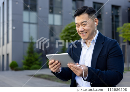 Close-up photo of a smiling young Asian man sitting outside in a suit and headphones, holding a tablet and talking on a video call. 113686208