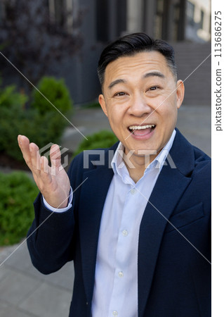 Happy Asian man standing outside in business suit and talking on video call gesturing with hands. Vertical photo. 113686275