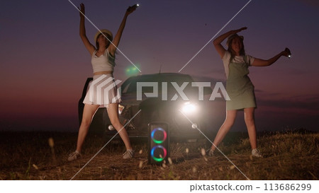 Women dancing at night in field near car. Happy cheerful satisfied girls friends having good time having party in late evening outdoors at nature near portable music speaker with ring rainbow lighting Women dancing at night in field near car. Happy cheerful satisfied girls friends having good time having party in late evening outdoors at nature near portable music speaker with ring rainbow lighting 113686299