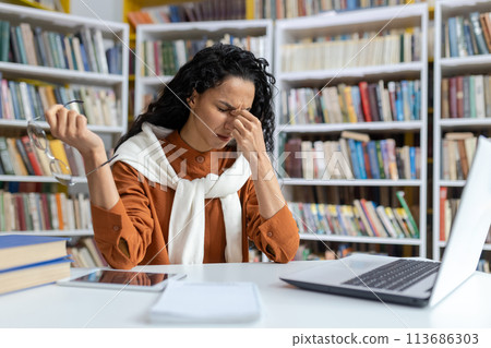 A Hispanic female student experiences stress and headache while working on her laptop in a library filled with books. 113686303