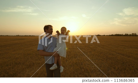 Father holding kid in arms in field at sunset. Happy cheery parent child kid strolling moving going across huge yellow meadow. Girl sits in man arms pointing with finger. Love fatherhood parenthood Father holding kid in arms in field at sunset. Happy cheery parent child kid strolling moving going across huge yellow meadow. Girl sits in man arms pointing with finger. Love fatherhood parenthood 113686365