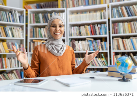 Confident and cheerful Muslim female teacher in a hijab smiling at the camera while standing in a library with shelves full of books. Confident and cheerful Muslim female teacher in a hijab smiling at the camera while standing in a library with shelves full of books. 113686371