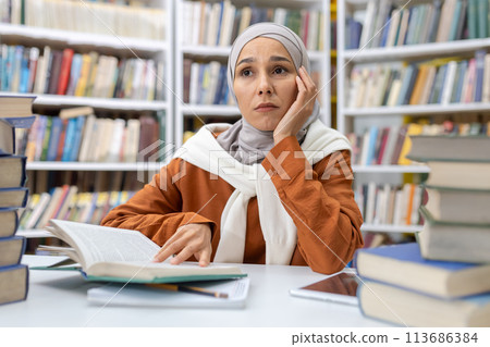 A thoughtful Muslim woman wearing a hijab sits amidst piles of books in a library, looking pensive and possibly overwhelmed by her studies or research. 113686384