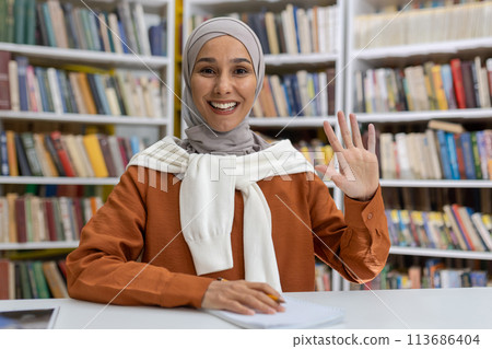 A joyful Muslim woman in a hijab waves at the camera while seated in a library, surrounded by books, radiating positivity and friendliness. 113686404