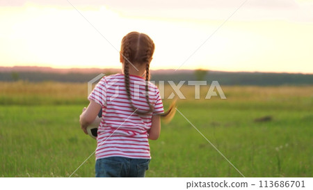 Active cute little girl kid running on sunset grass field with football ball back view closeup. Funny female child having fun playing with soccer sports enjoy happy childhood outdoor at summer meadow 113686701