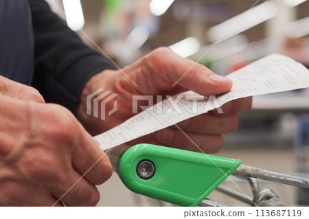 The hands of an unrecognizable older man hold a sales receipt next to a grocery cart in a supermarket. The hands of an unrecognizable older man hold a sales receipt next to a grocery cart in a supermarket. 113687119