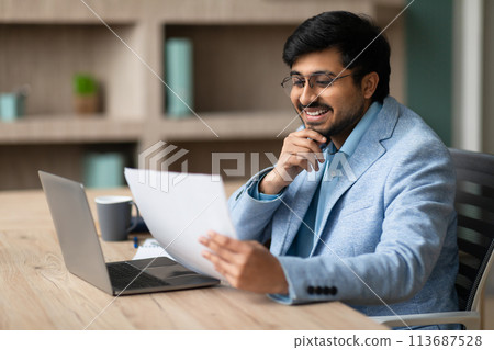 Contented Indian businessman looks through papers engrossed in work indoor 113687528