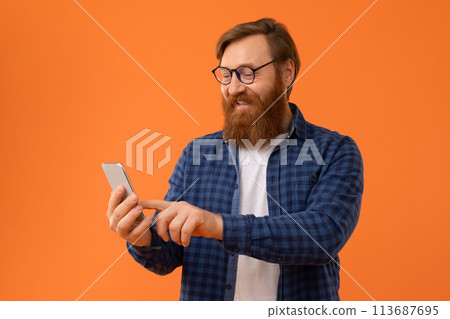 redhaired Caucasian man with beard uses his smartphone, orange backdrop 113687695