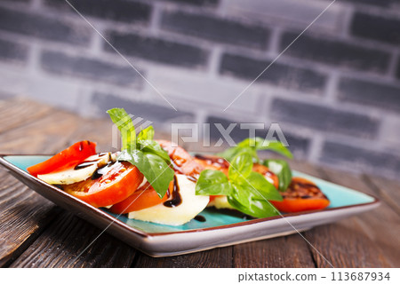 Italian caprese salad with sliced tomatoes, mozzarella, basil, olive oil on wooden background. Top view. Italian caprese salad with sliced tomatoes, mozzarella, basil, olive oil on wooden background. Top view. 113687934
