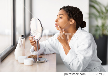 Concerned young black woman in white bathrobe examining herpes outbreak on lip Concerned young black woman in white bathrobe examining herpes outbreak on lip 113687936