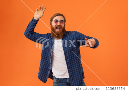 Happy redhaired bearded guy in sunglasses pointing over orange background 113688354