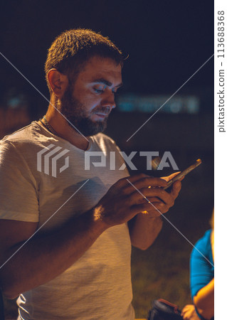 A brutal man writes a message on his phone at night in a city park. A handsome man with a phone is walking through a city park on a summer night. Concept of a man talking on the phone in the park. 113688368