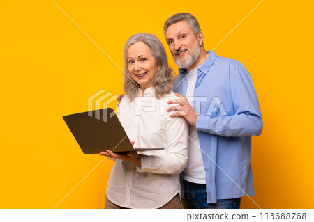 Smiling elderly husband and wife standing with computer, yellow studio 113688768