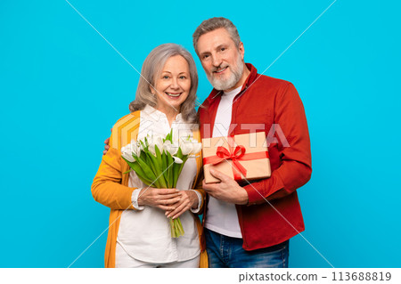 Happy senior couple posing with present and bouquet in studio Happy senior couple posing with present and bouquet in studio 113688819