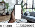 Augmented reality in interior design mockup. Close-up view of a black hand holding smartphone with transparent blank screen, ideal for digital AR design 113689005
