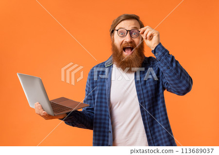 Excited redhaired bearded programmer guy holds laptop over orange background 113689037