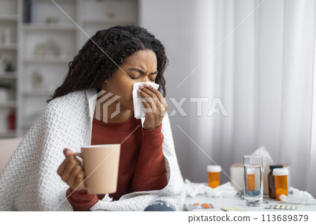 Black woman suffering cold, blowing her nose and holding cup of tea 113689879