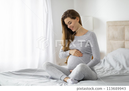 Pregnant woman sitting on bed holding belly 113689943