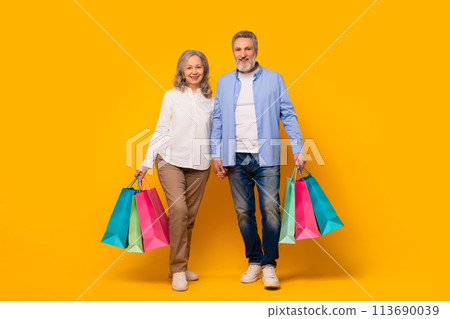 Joyful elderly couple holds shopping bags against yellow studio backdrop 113690039