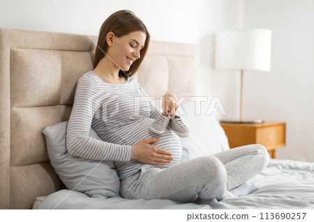Pregnant woman holding baby shoes on bed 113690257
