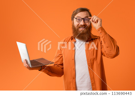 Cheerful gingerhead bearded man works on laptop on orange backdrop Cheerful gingerhead bearded man works on laptop on orange backdrop 113690443