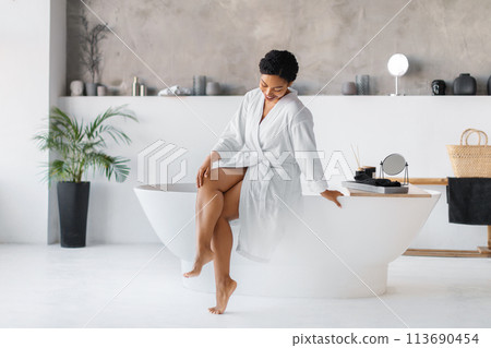 Black woman wearing bathrobe sitting on bathtub and touching her beautiful legs 113690454