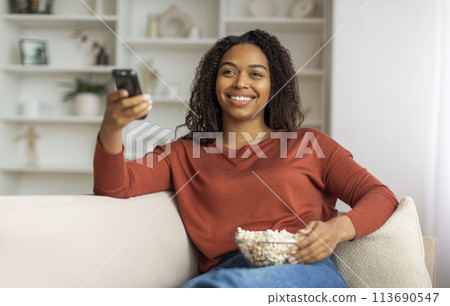 Smiling Black Woman Watching TV and Holding Remote at Home 113690547