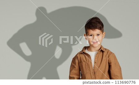 A happy young boy in a brown jacket smiles brightly, standing in front of a gray backdrop with a shadow 113690776