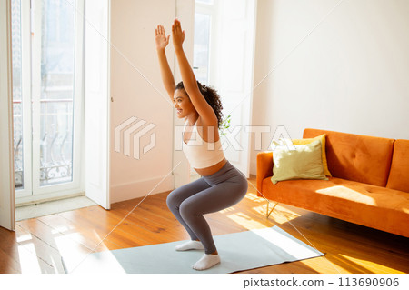 Fit active lady in sportswear squatting with raised arms indoor 113690906