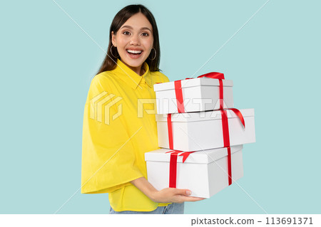Excited young woman holding stylish white gifts with bold red ribbons at blue background Excited young woman holding stylish white gifts with bold red ribbons at blue background 113691371